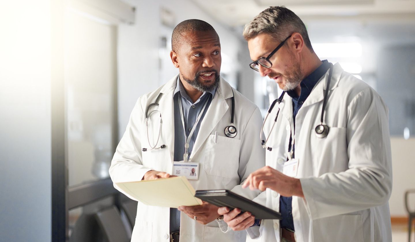 Two doctors in white coats in a hospital corridor, discussing a patient's file and a tablet.